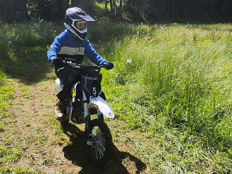 Un jeune garçon seul sur sa mini moto roulant doucement sur un sentier bordé de hautes herbes.