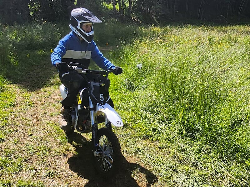 Un jeune garçon seul sur sa mini moto roulant doucement sur un sentier bordé de hautes herbes.