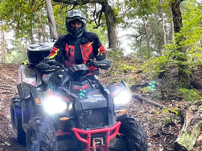 Conducteur en tenue rouge et noire sur un quad Polaris, roulant dans une forêt sur un chemin boueux avec les phares allumés.