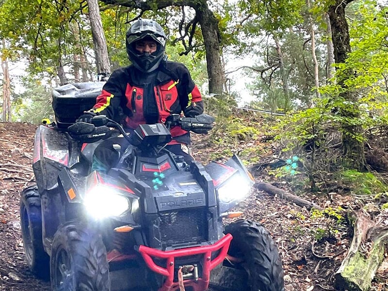 Conducteur en tenue rouge et noire sur un quad Polaris, roulant dans une forêt sur un chemin boueux avec les phares allumés.