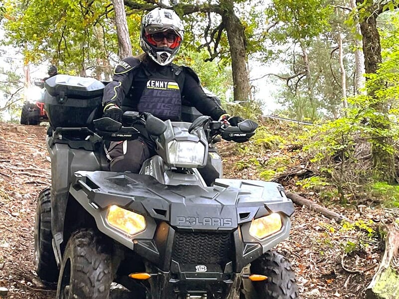 Personne équipée d’un casque et d’une veste Kenny noire conduisant un quad Polaris sur un sentier forestier en descente.