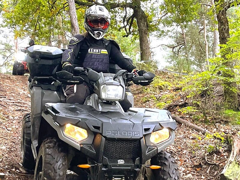 Personne équipée d’un casque et d’une veste Kenny noire conduisant un quad Polaris sur un sentier forestier en descente.