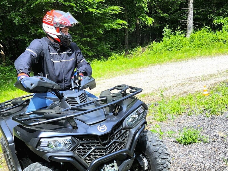 Homme casqué sur un quad noir en pleine manœuvre sur un terrain dégagé près d’une forêt.