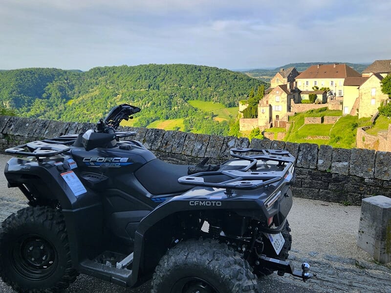 Quad CFMOTO stationné sur un promontoire en pierre avec vue panoramique sur une vallée verdoyante et un village médiéval.