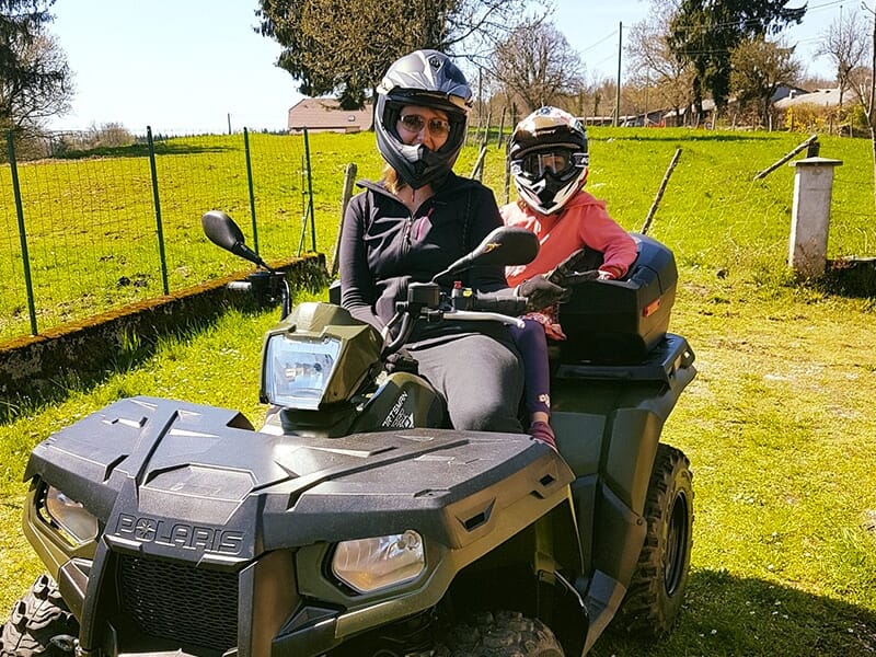 Femme et enfant portant des casques, assis ensemble sur un quad Polaris dans un jardin ensoleillé à la campagne.