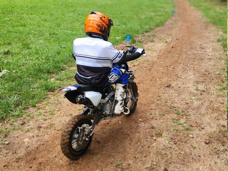 Pilote enfant en casque orange et tenue blanche, vu de dos, conduisant une moto lors d'un stage motocross enfant près de Lons-le-Saunier.
