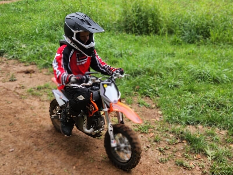 Enfant en tenue rouge et noire avec casque noir, conduisant une petite moto orange lors d'un stage motocross enfant près de Lons-le-Saunier.
