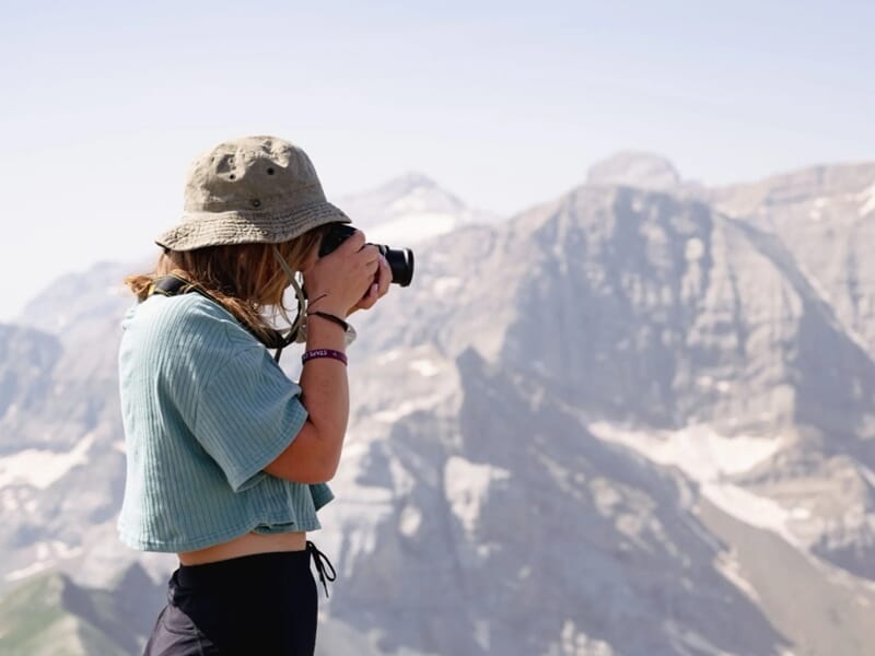 Jeune femme photographiant un paysage alpin avec un reflex, vêtue d’un bob et d’un haut court.