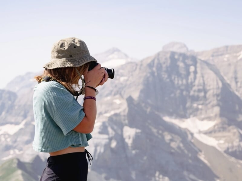 Jeune femme photographiant un paysage alpin avec un reflex, vêtue d’un bob et d’un haut court.