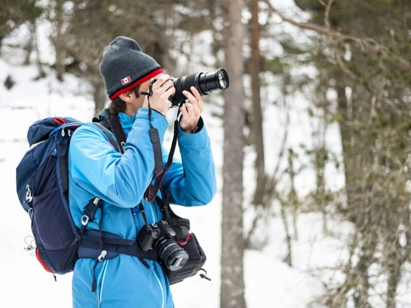 Photographe en manteau bleu et bonnet, équipé de deux appareils photo dans une forêt enneigée.