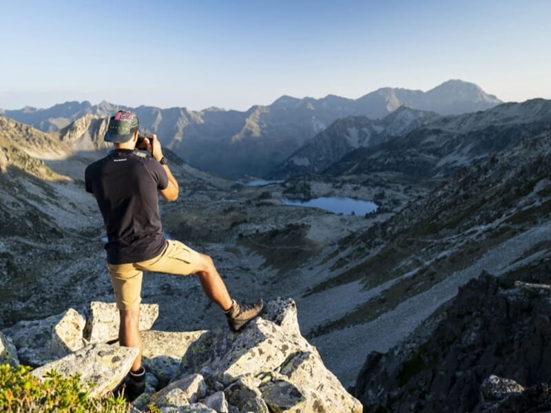 Photographe en tenue de randonnée capturant un panorama de montagnes et de lacs depuis un sommet rocheux