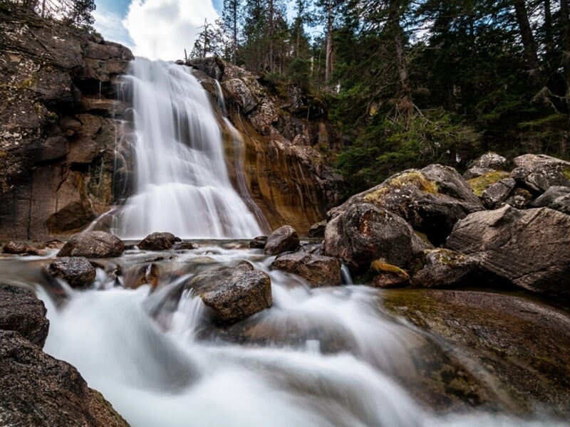 Cascade en forêt photographiée en pose longue, avec des rochers au premier plan