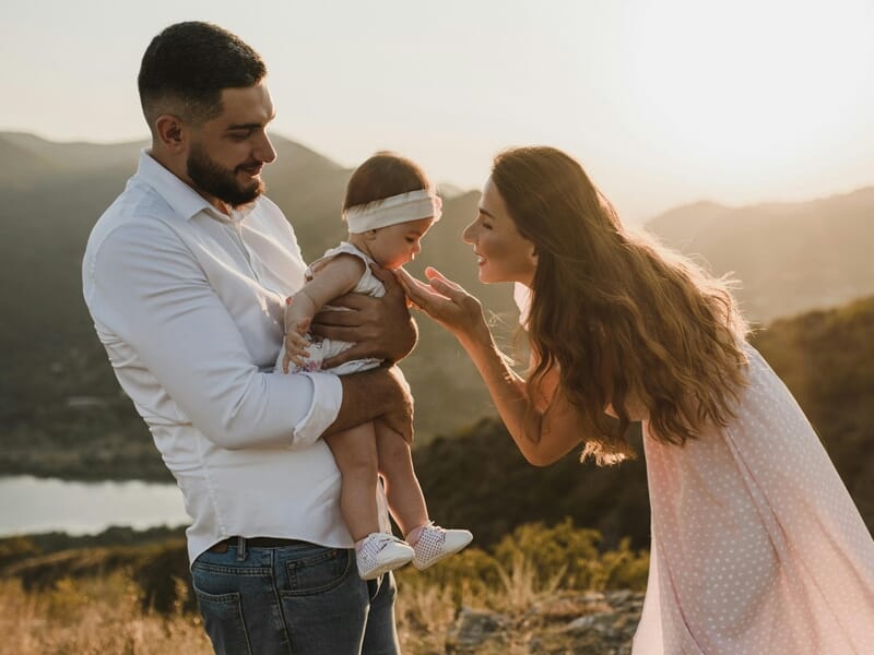 Un père tient son bébé dans les bras pendant que la mère lui caresse le visage au coucher du soleil en montagne.