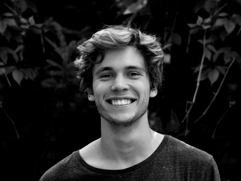 Portrait en noir et blanc d’un homme souriant debout devant un fond de feuillage.