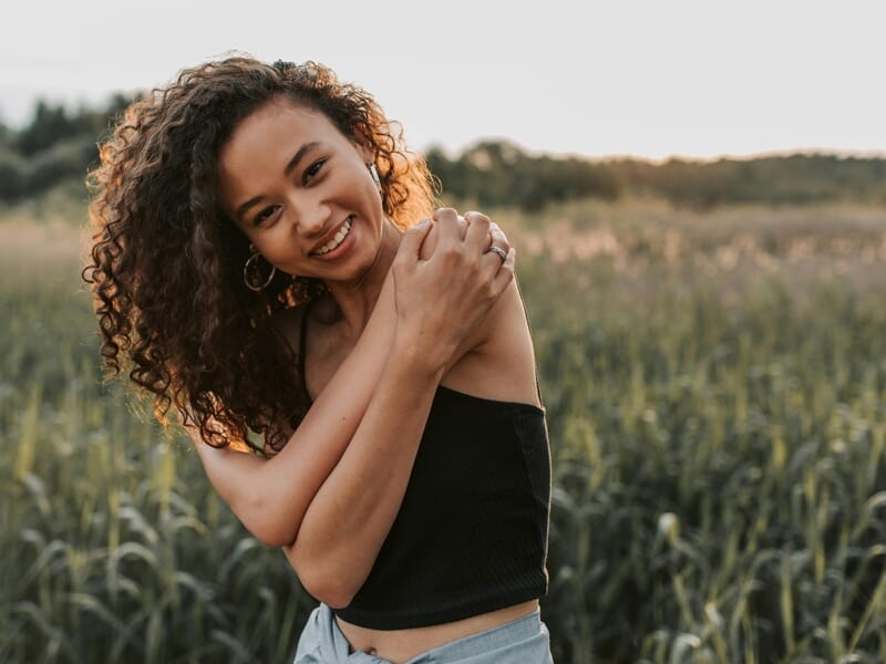 Jeune femme souriante avec les bras croisés sur les épaules, debout dans un champ au coucher du soleil.