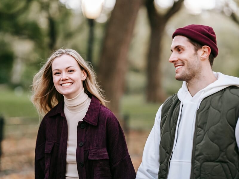 Jeune couple souriant marchant dans un parc, la femme rit tandis que l’homme la regarde.