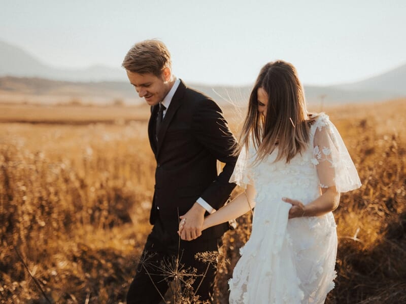Couple main dans la main courant dans un champ, la femme porte une robe de mariée blanche.