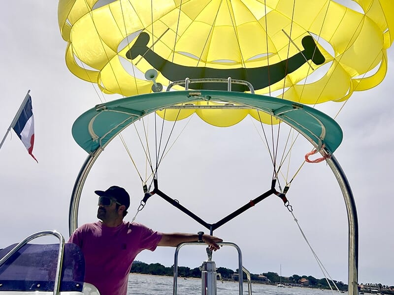 Homme portant une casquette et un t-shirt rose pilotant un bateau de parachute ascensionnel, avec un parachute jaune décoré d’un smiley au-dessus.