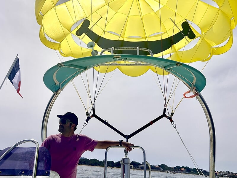 Homme portant une casquette et un t-shirt rose pilotant un bateau de parachute ascensionnel, avec un parachute jaune décoré d’un smiley au-dessus.