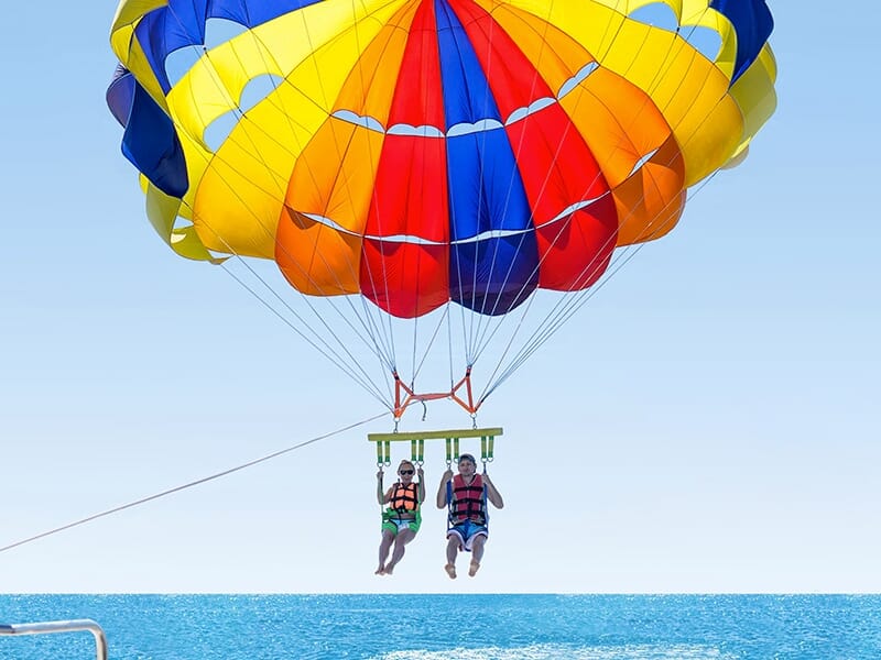 Deux personnes en gilet de sauvetage en plein vol en parachute ascensionnel au-dessus de la mer, attachées à un parachute coloré.