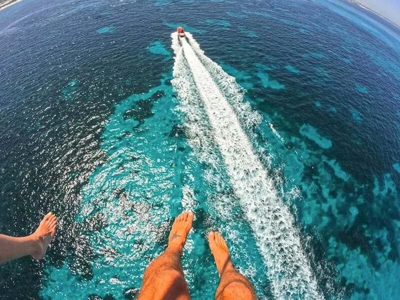 Vue depuis les airs de deux personnes en parachute ascensionnel au-dessus d’une mer turquoise, avec un bateau à moteur traçant une longue traînée blanche.