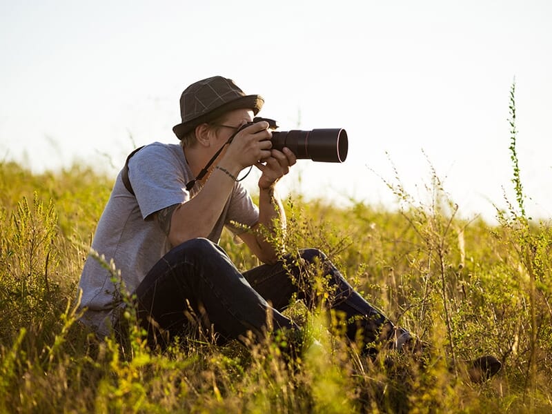 Photographe assis dans l’herbe prenant une photo avec un téléobjectif, en pleine nature.