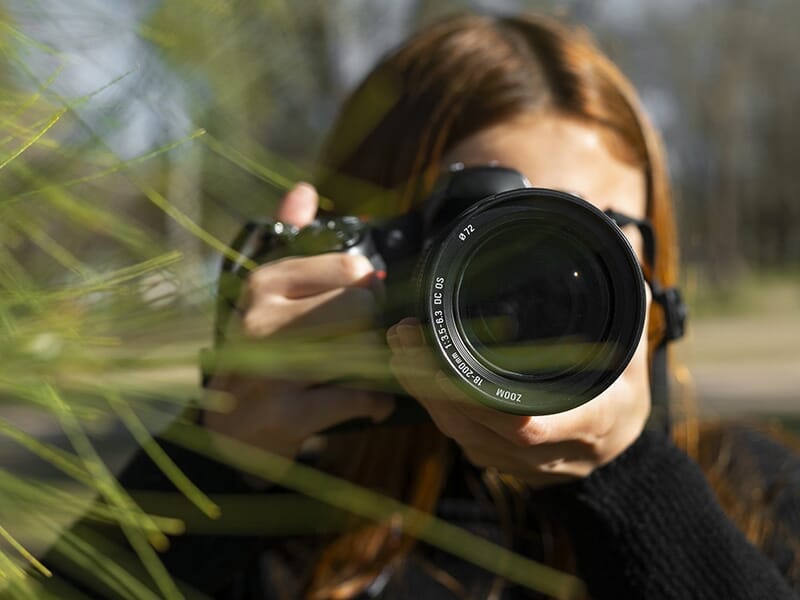 Jeune femme tenant un appareil photo avec objectif zoom, camouflée partiellement par des branches.