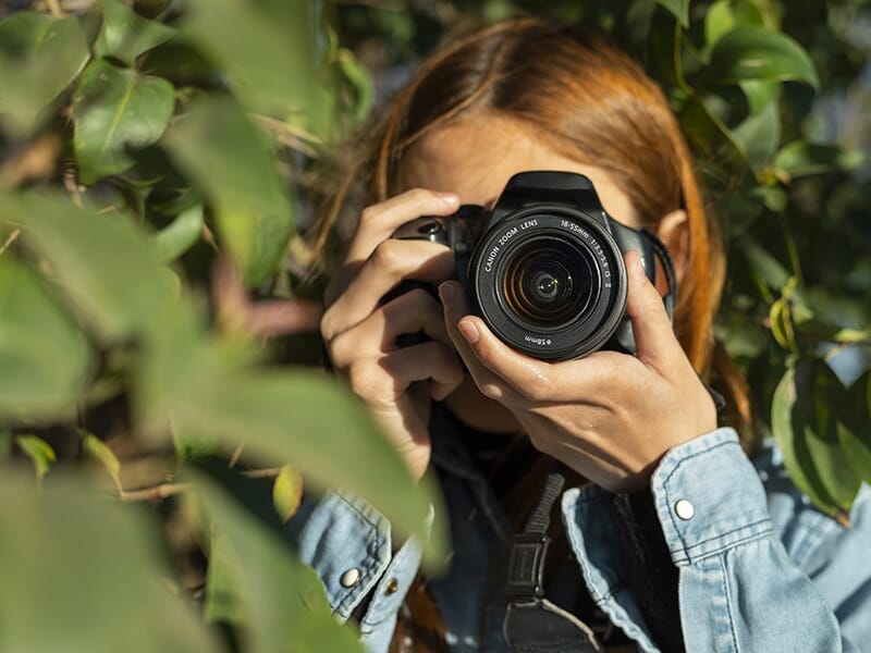 Jeune photographe tenant un reflex numérique au milieu de feuillage vert.