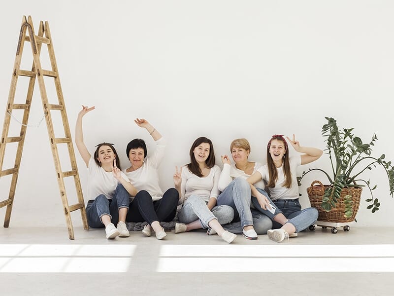 Groupe de cinq femmes assises au sol contre un mur blanc, posant joyeusement dans un studio lumineux avec une échelle en bois et une plante verte.
