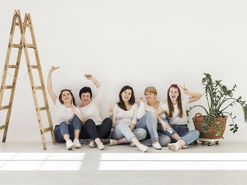 Groupe de cinq femmes assises au sol contre un mur blanc, posant joyeusement dans un studio lumineux avec une échelle en bois et une plante verte.