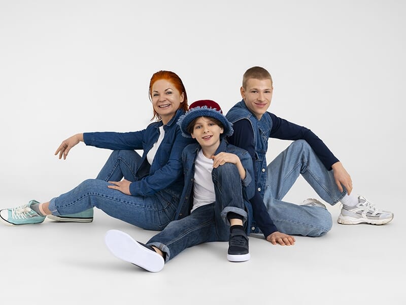 Portrait en studio d’une famille souriante composée d’une mère et de deux enfants, tous habillés en jean.