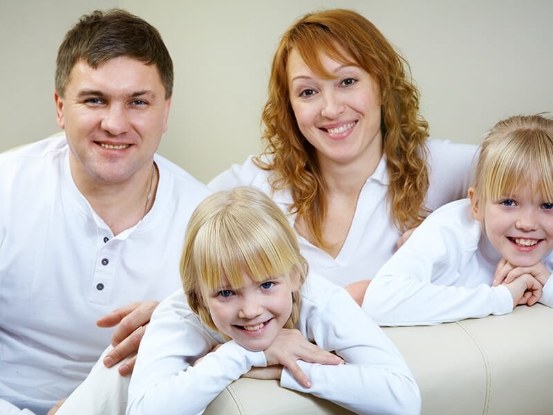Portrait chaleureux d’une famille à quatre, avec les parents et deux jeunes filles blondes, allongés sur un canapé clair.