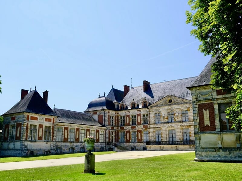 Château ancien aux façades de pierre et briques rouges, sur pelouse verte, sous ciel bleu sans nuage.