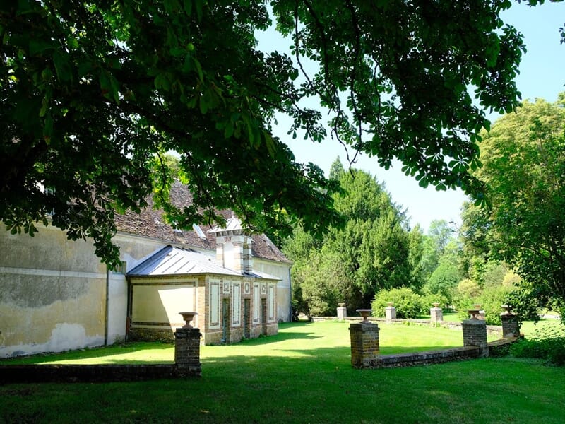 Bâtiment bas avec murs anciens et toiture pentue, entouré de verdure dense et ombragée.