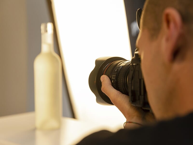Photographe en gros plan prenant une bouteille en photo dans un studio avec éclairage diffus à l’arrière-plan.