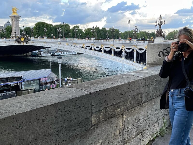 Femme souriante réglant son appareil photo sur trépied au coucher du soleil, sur le pont Alexandre III, avec la Seine en arrière-plan.
