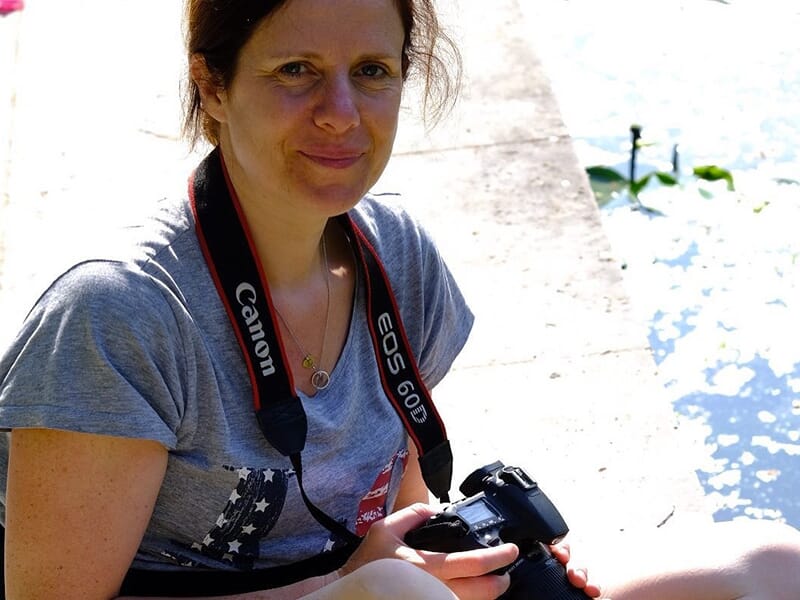 Femme assise en extérieur, tenant un appareil photo Canon EOS 60D, avec une sangle autour du cou, près d'un plan d'eau ensoleillé