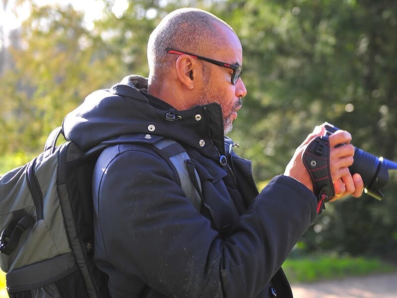 Un homme examine son appareil photo reflex en pleine nature, portant un sac à dos et des lunettes de soleil.