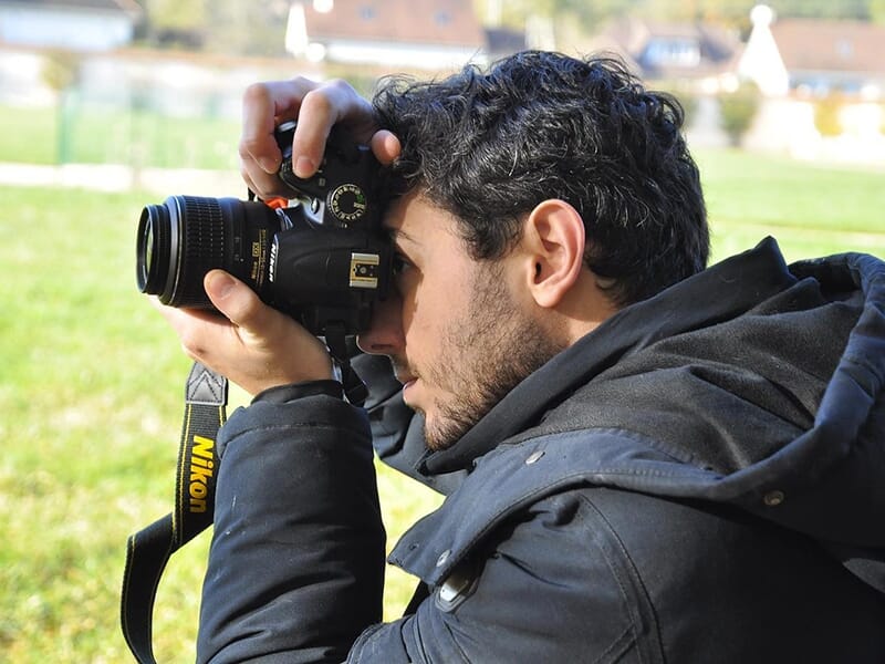 Un homme photographie un paysage avec un appareil Nikon, en extérieur, vêtu d’une veste noire.