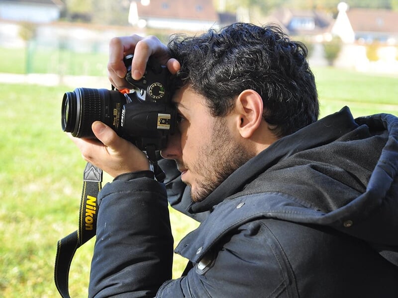 Un homme photographie un paysage avec un appareil Nikon, en extérieur, vêtu d’une veste noire.