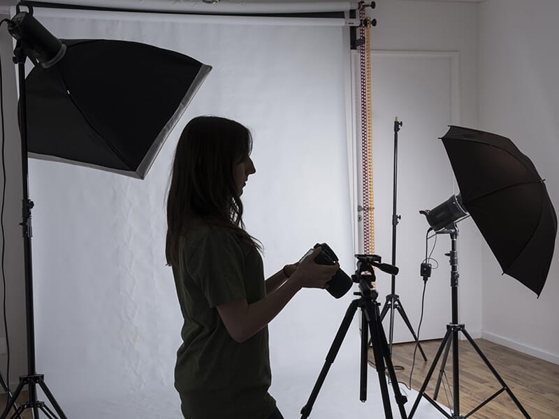 Photographe en contre-jour réglant son appareil dans un studio équipé de softbox et d’un fond blanc.