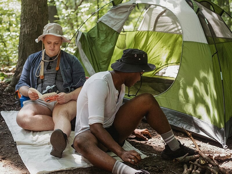 Deux campeurs assis devant leur tente, en pleine forêt, en train de lire et d’allumer un feu.
