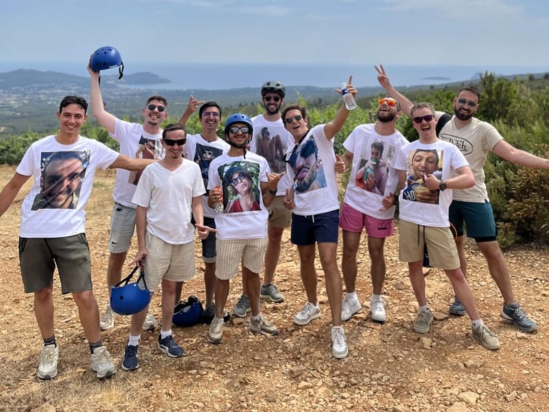 Groupe de dix hommes souriants en t-shirts blancs avec photos, posant en plein air avec un casque bleu en main.