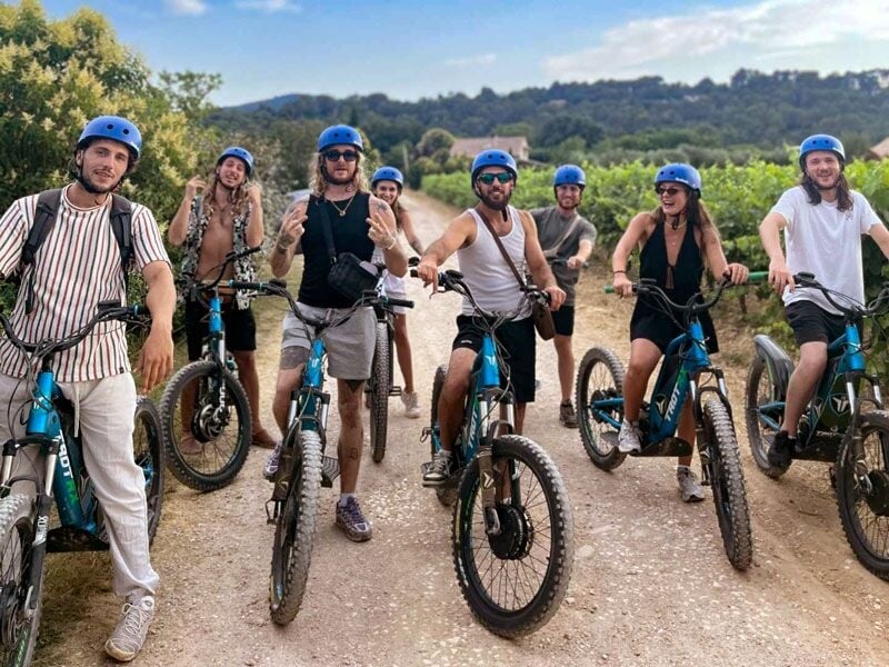 Groupe de sept personnes souriantes portant des casques bleus, posant avec leurs vélos électriques sur un chemin en pleine nature.