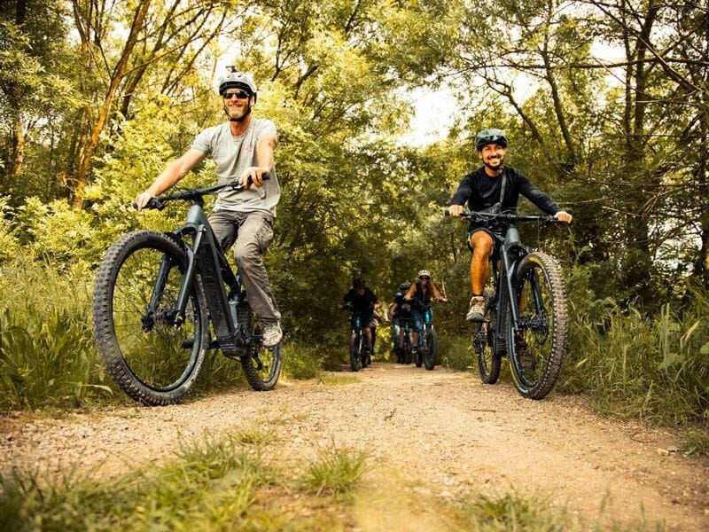 Deux hommes souriants font du vélo électrique sur un chemin forestier entouré d'arbres verts en été.