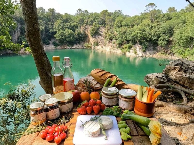 Table en plein air avec assortiment de fruits, légumes, pains et boissons au bord d'un lac turquoise.