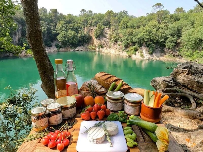 Table en plein air avec assortiment de fruits, légumes, pains et boissons au bord d'un lac turquoise.