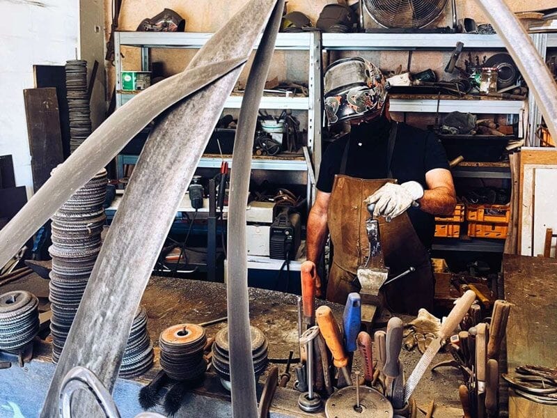 Artisan forgeron portant un tablier et un casque de protection travaillant dans un atelier avec outils.