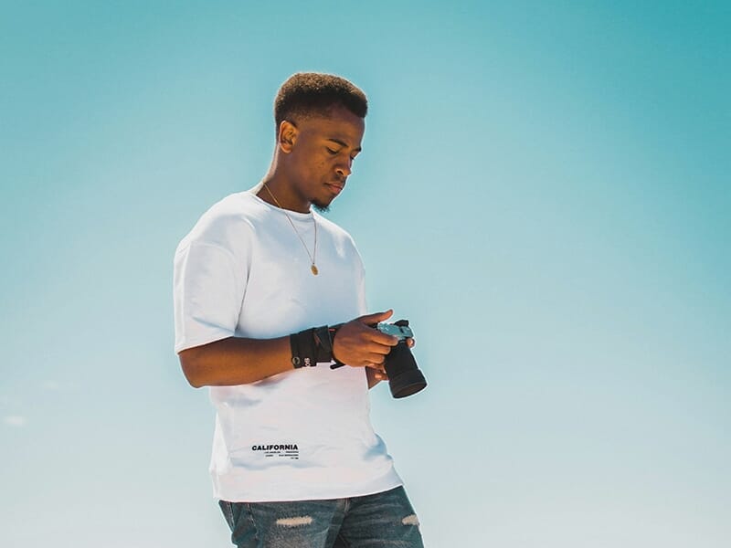 Jeune homme en t-shirt blanc observant son appareil photo sous un ciel bleu clair.