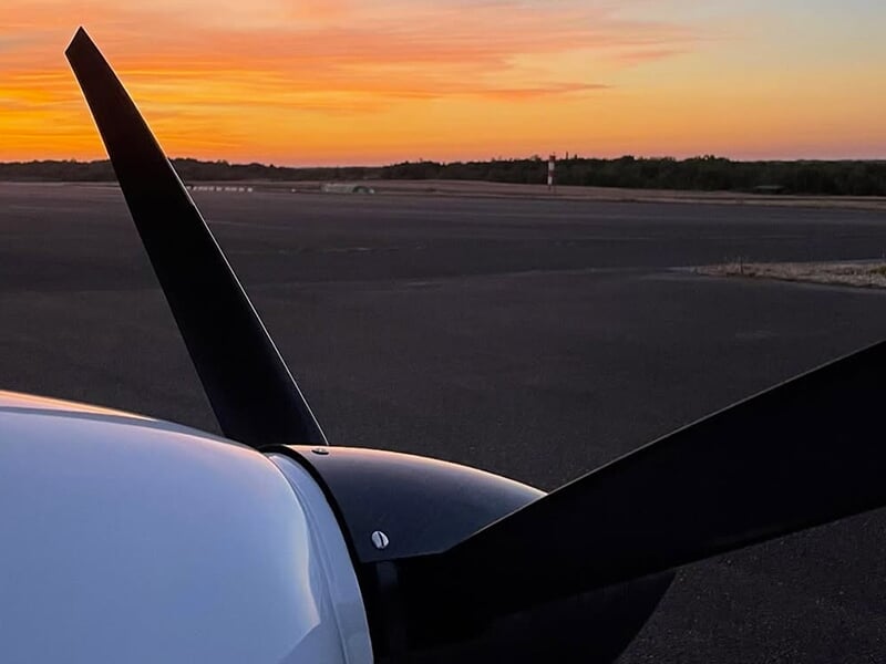 Hélice d’un avion léger devant un ciel de coucher de soleil sur une piste d’aéroport.